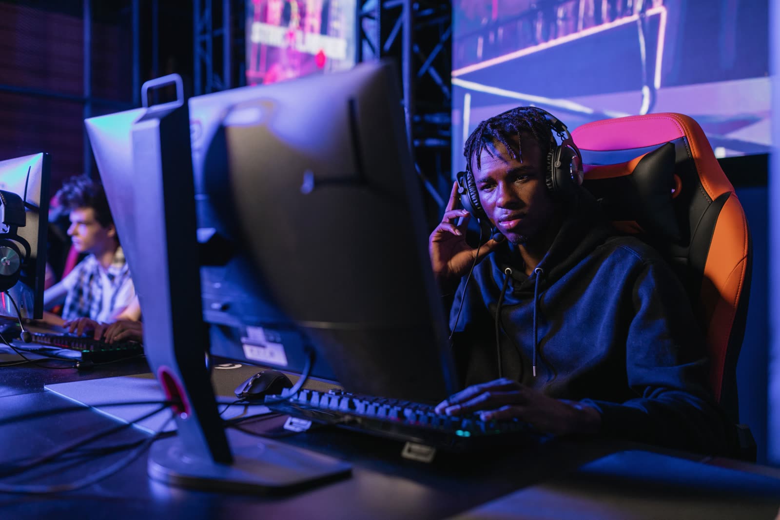 A player in a headset at a club station, hand on the keyboard, with another player at the next monitor under blue-violet lighting